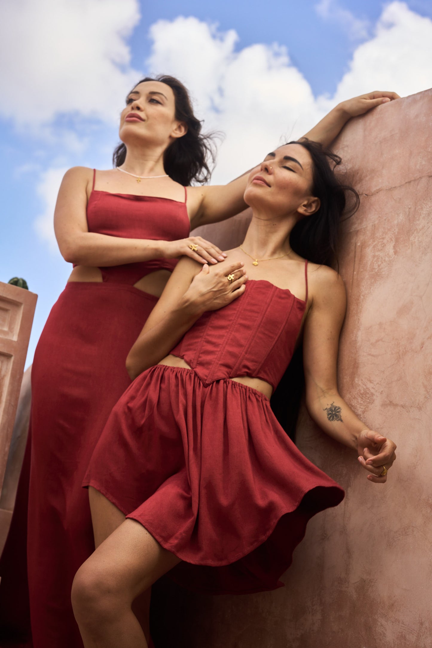 Two women in red dresses together against a pink wall with a blue sky.