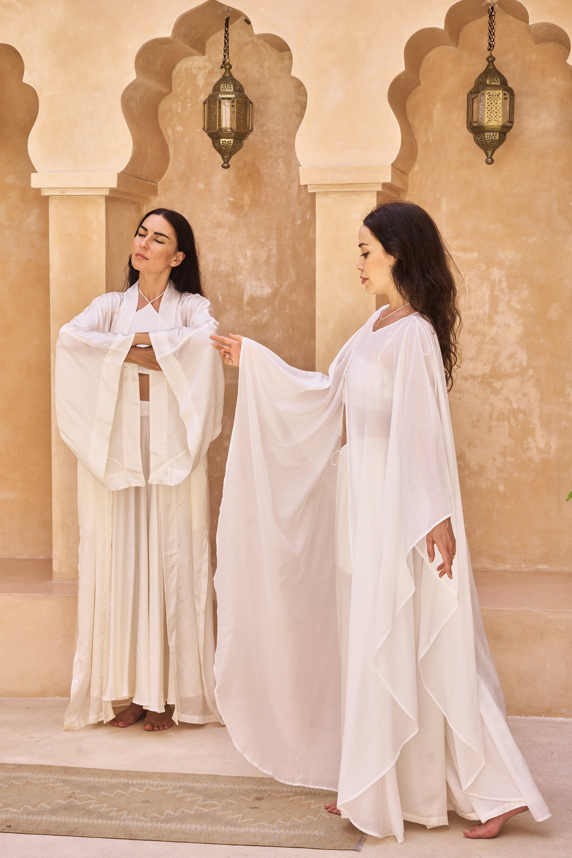Two women in white flowing kimono robes standing in front of a beige wall with arched doorways.