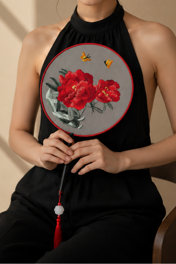 A woman seated on a wooden chair wearing a black sleeveless halter top and black pants, holding a round fan with a red floral design. The background is a warm beige wall with soft window shadow patterns.