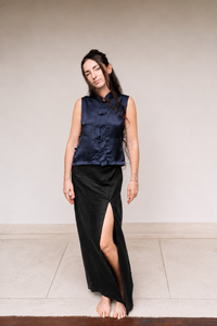 A woman stands barefoot against a plain beige wall wearing a sleeveless navy satin vest with traditional collar and button details, paired with a long black skirt featuring a high side slit.