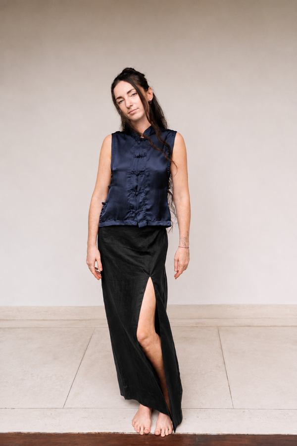A woman stands barefoot against a plain beige wall wearing a sleeveless navy satin vest with traditional collar and button details, paired with a long black skirt featuring a high side slit.