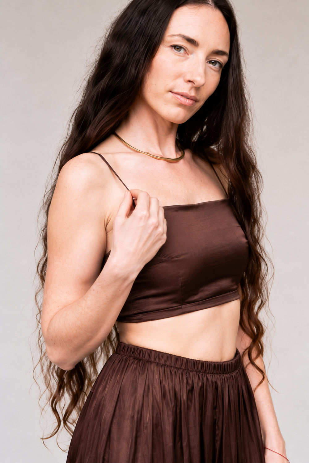 A woman with long, wavy dark brown hair wearing a matching dark brown satin crop top and high-waisted pleated skirt, photographed against a neutral light gray background.