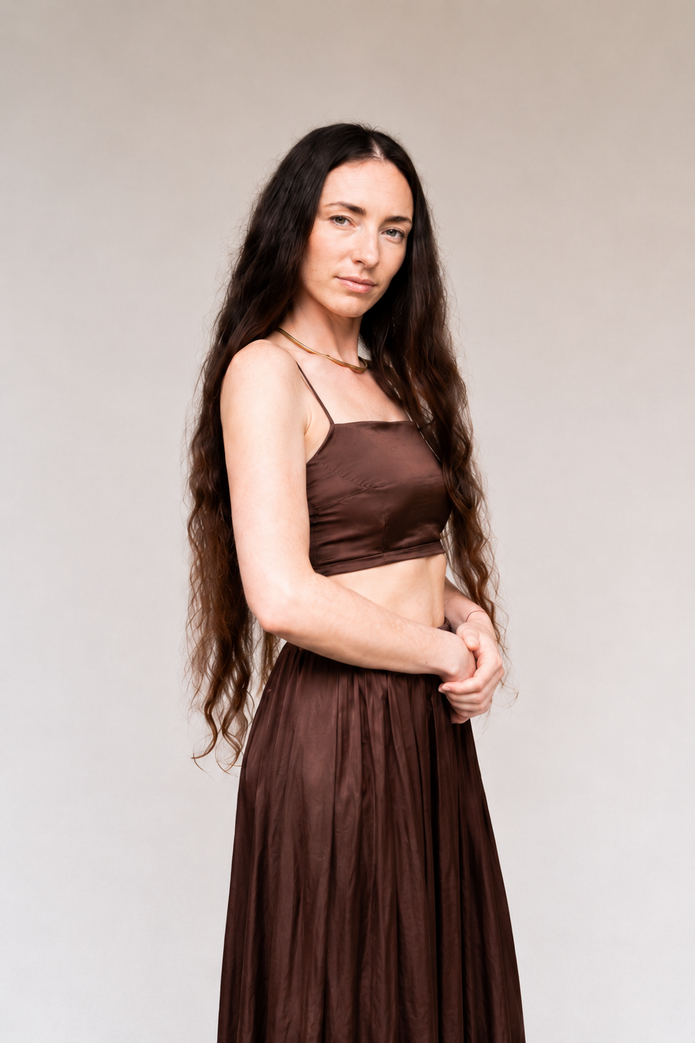 A woman with long, wavy dark brown hair is wearing a matching two-piece brown satin and chiffon set, consisting of a spaghetti-strap crop top and a high-waisted pleated maxi skirt, against a plain light gray studio background.
