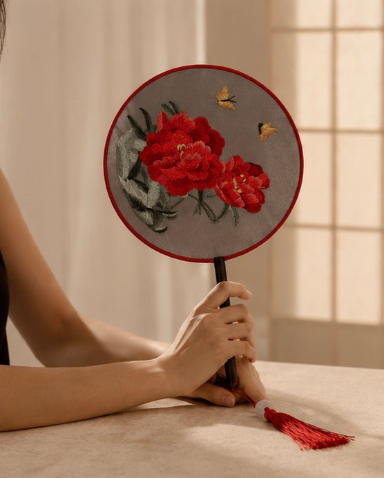 A person holds a round embroidered fan with red peony flowers, green leaves, and two small butterflies on a gray fabric background. The fan has a red border, a black handle, and a red tassel, set against a warm beige background with soft window light.