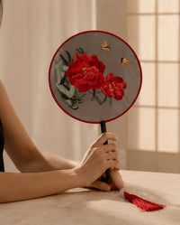 A person holds a round embroidered fan with red peony flowers, green leaves, and two small butterflies on a gray fabric background. The fan has a red border, a black handle, and a red tassel, set against a warm beige background with soft window light.