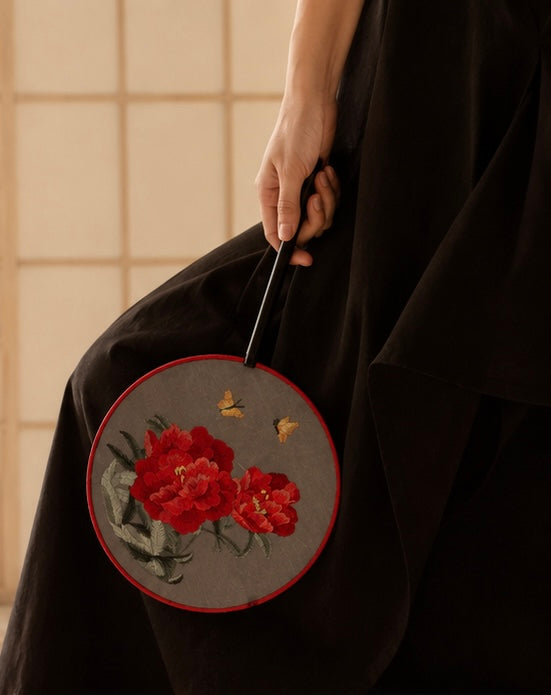 A close-up of a person in a black kimono holding a round embroidered fan. The fan has a gray fabric body with red embroidered flowers, green leaves, and two small yellow butterflies, framed by a red border.
