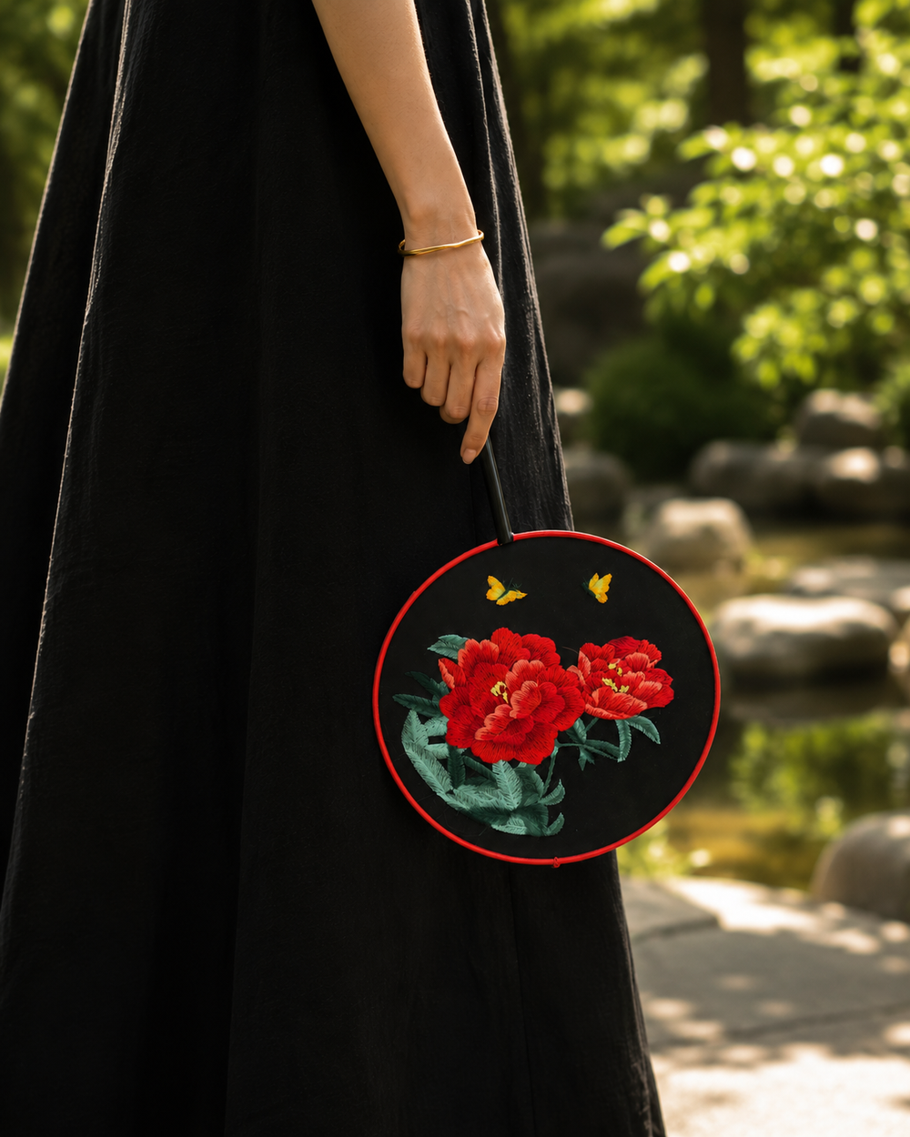 A person in a long black dress holds a round black handbag with embroidered red flowers, green leaves, and small yellow butterflies, photographed outdoors near a garden pond.
