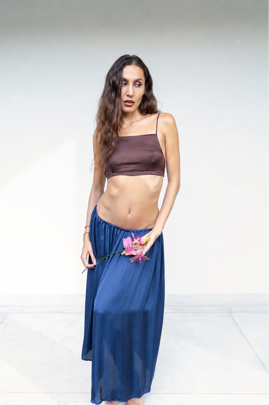 A woman stands barefoot against a plain white studio background, wearing a dark brown Rui top and a flowing midnight blue lin skirt. She is holding a pink flower.