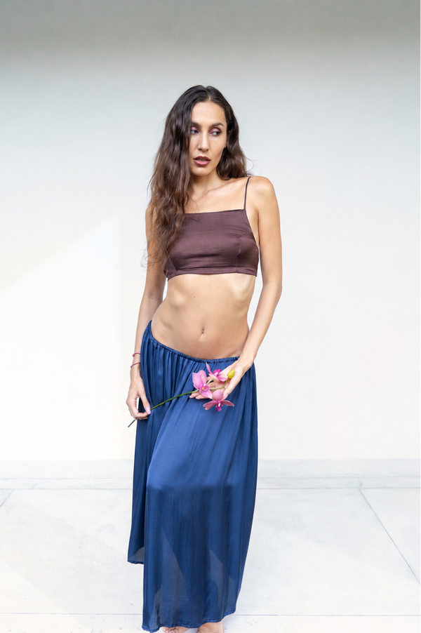 A woman stands barefoot against a plain white studio background, wearing a dark brown Rui top and a flowing midnight blue lin skirt. She is holding a pink flower.