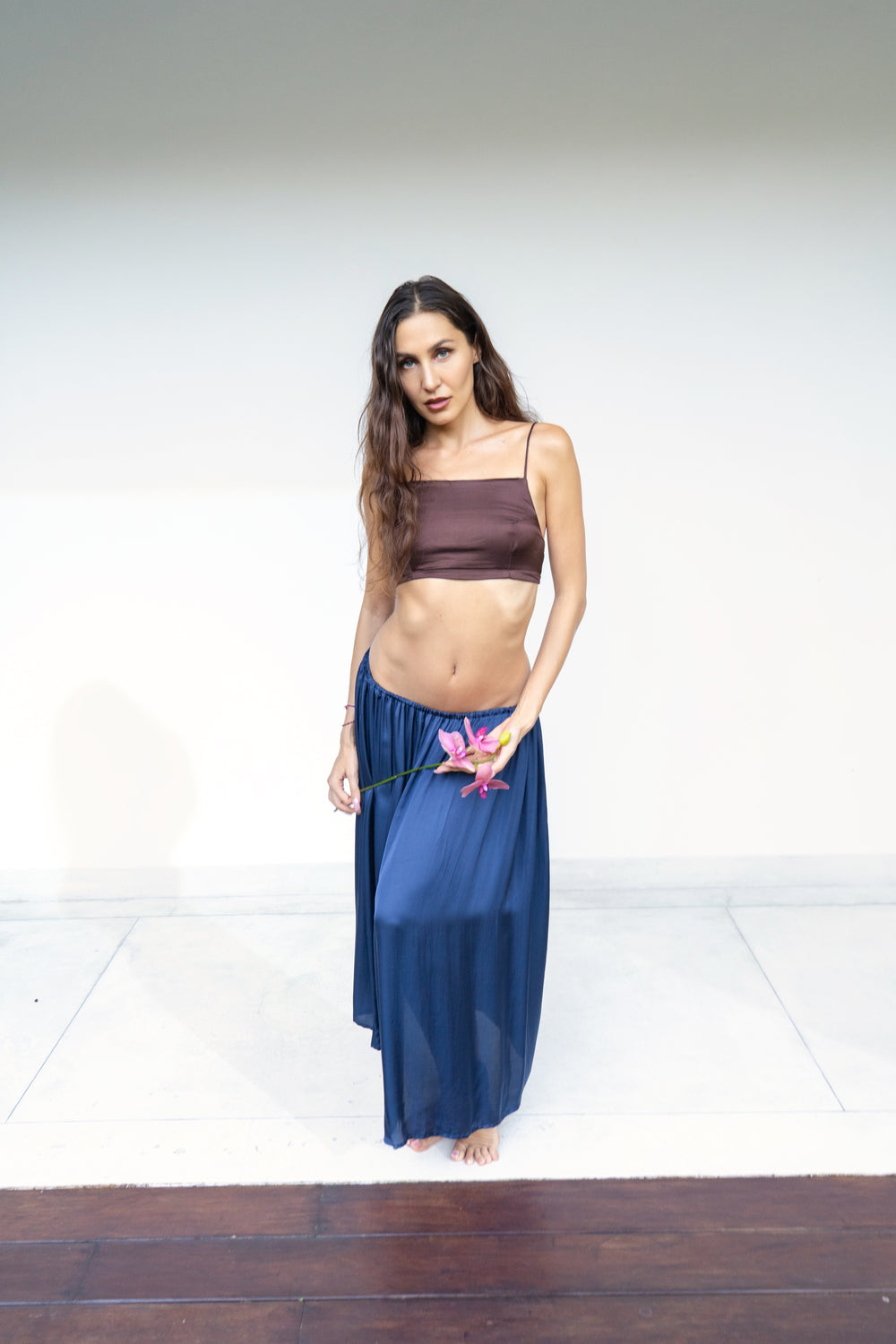 A woman stands barefoot against a plain white studio background, wearing a dark brown Rui top and a flowing midnight blue lin skirt. She is holding a pink flower.