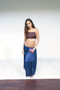 A woman stands barefoot against a plain white studio background, wearing a dark brown Rui top and a flowing midnight blue lin skirt. She is holding a pink flower.