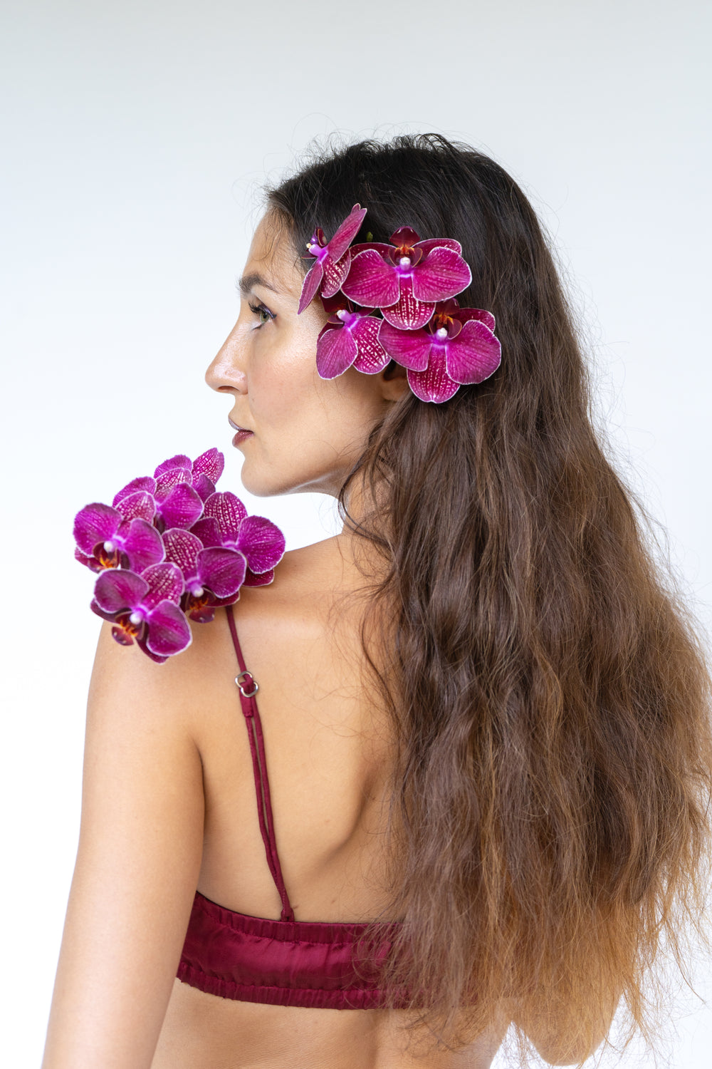 A woman with long wavy brown hair is shown in profile against a light gray studio background. She is wearing bright magenta-purple orchid flower hair clips, one attached to her hair and one near her shoulder, and a matching deep burgundy satin bralette.