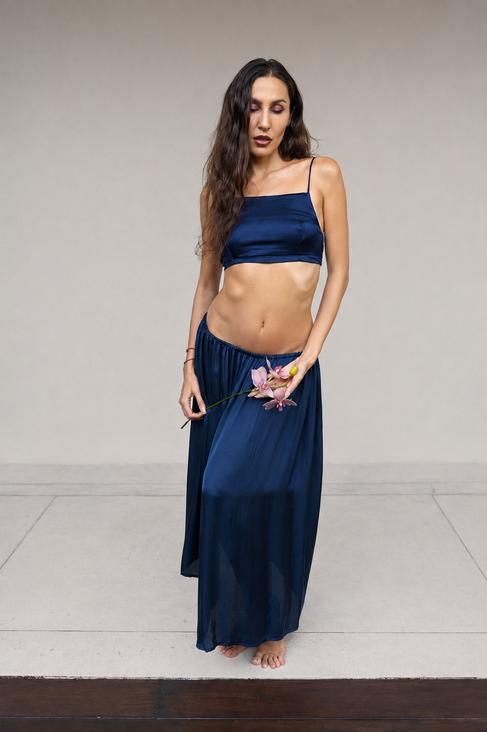 A woman modeling a navy blue silk two-piece set consisting of a Rui top and a Lin skirt, standing against a neutral light gray studio background.