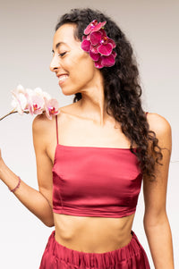 A woman with long curly dark hair wearing a matching maroon silk crop top and pants, holding a pink orchid flower. She has a large pink orchid flower tucked into her hair.