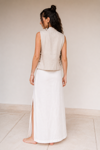 A woman shown from the back wearing a sleeveless light beige vest and a long linen white skirt with a side slit, standing barefoot against a plain light beige wall and floor.