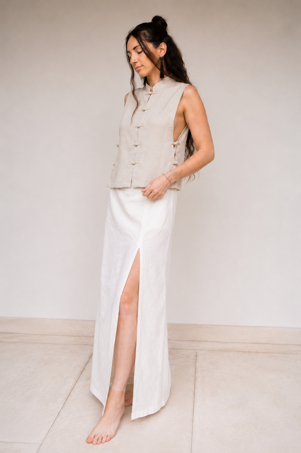 A woman stands barefoot in a minimalist studio setting, wearing a sleeveless light beige vest with Chinese inspired buttons and a long linen white skirt featuring a high side slit.