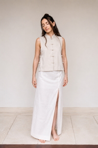 A woman stands barefoot in a minimalist studio setting, wearing a sleeveless light beige vest with traditional button details and a long white skirt with a high side slit.